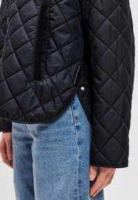 Čierna prešívaná bunda s matným povrchom, bočným zipsovým vreckom a zaobleným lemom. Doplnok k svetlomodrým džínsom. Textúrovaný, ľahký materiál.