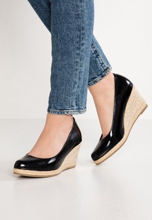 Sorte laklæder kilehøjhæle med fletplatform af jutegarn, båret med opkrogede blå jeans på lys gulv.