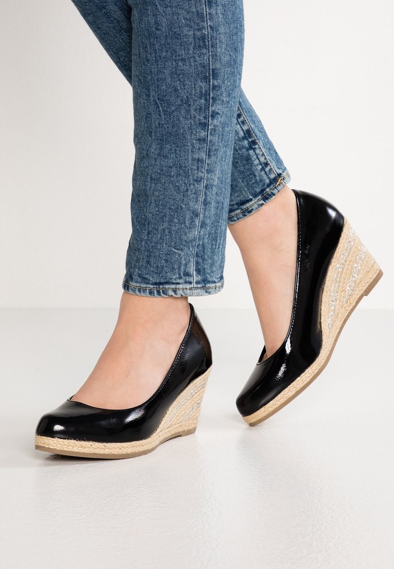 Escarpins compensés en cuir verni noir avec plateforme en jute tressée, portés avec un jean bleu retroussé sur un sol clair.