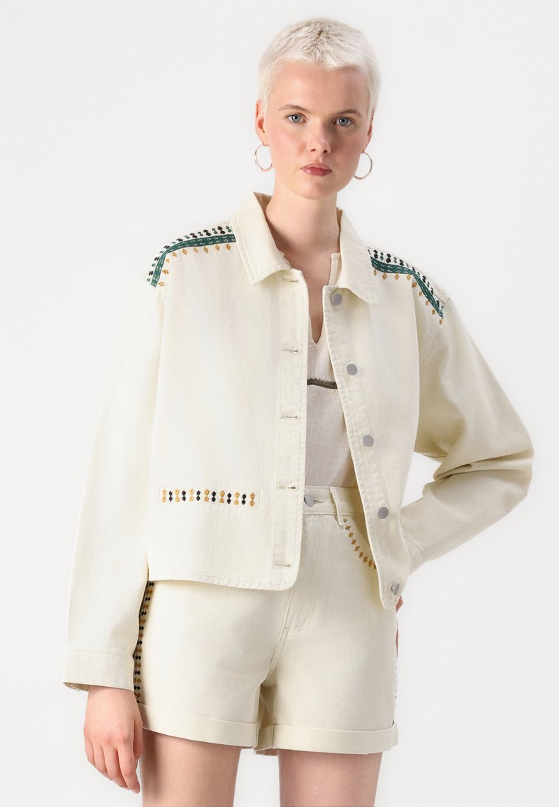 Veste en denim crème avec broderie bleue et dorée sur les épaules, dotée d'un col et d'une fermeture à boutons. Associée à un short assorti.