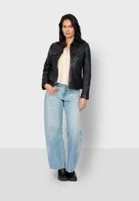 Woman with long dark hair wearing a black leather jacket, cream top, light blue wide-leg jeans, and black boots standing against a plain background.