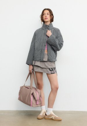 Jonge vrouw draagt een grijze gewatteerde jas, een gelaagde rok, witte sokken, beige sneakers en draagt een beige weekendtas met roze accenten.