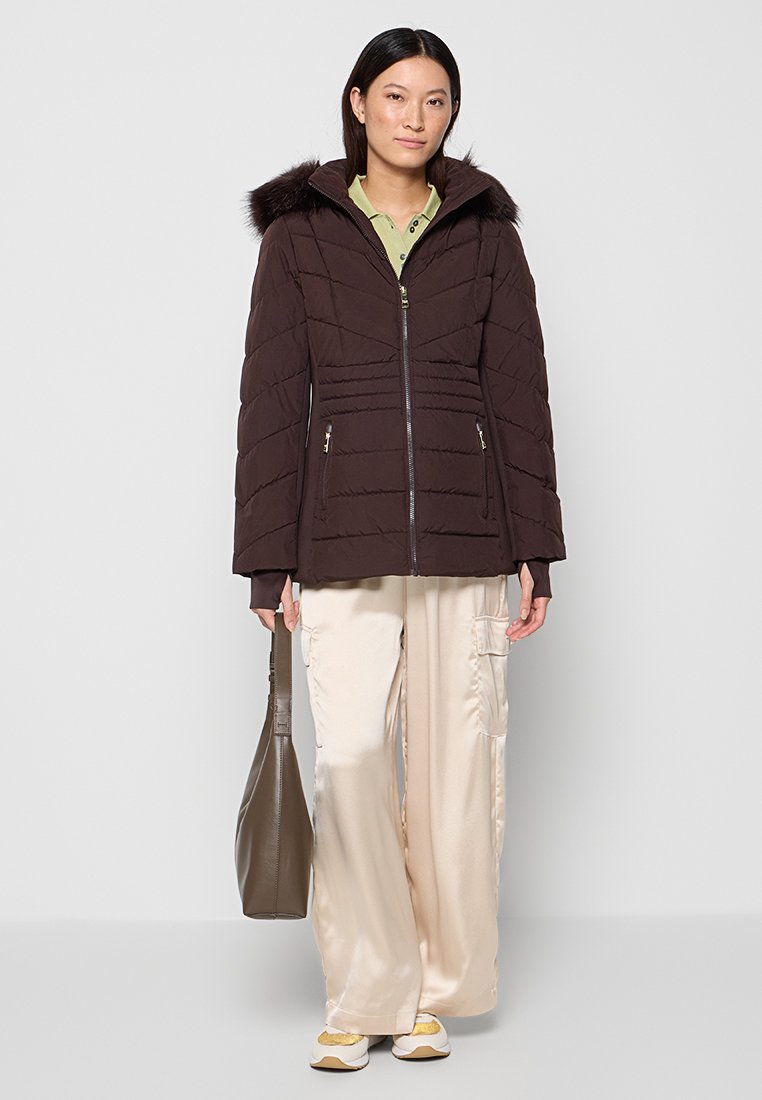 Vrouw die staat en een donkerbruine gebroken jasje met een bontkraag draagt, beige wijde broek, witte sneakers, en een grijze handtas vasthoudt.