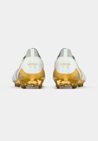Ποδοσφαιρικά παπούτσια με λευκό πάνω μέρος, γκρι λεπτομέρειες και σόλα χρυσού χρώματος. Διαθέτουν υφή και μυτερά καρφιά για καλύτερη πρόσφυση.