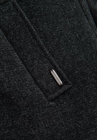 Mörkgrått tyg med ett strukturerat mönster, med en liten silveretikett märkt "QUINTESSENCE." Strukturerad design med fickdetalj.