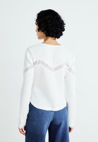 Hvit langermet genser med et teksturert strikkemønster og blonder i chevron-design på baksiden. Kombinert med blå denimbukser.