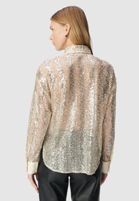 Blusa trasparente decorata con paillettes, base beige chiaro e dettagli argento, con collo a camicia e maniche lunghe con polsini crema.