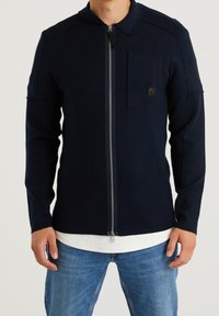 Marinblå dragkedjetröja med krage, som har en bröstficka och texturerad tyg. Anmärkningsvärda detaljer inkluderar en silverfärgad dragkedja och logotypdetalj.