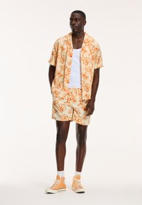 Korte mouwen bloemenoverhemd en bijpassende shorts, beide in lichtbeige met oranje patronen. Accessoires zijn oranje sneakers en een wit mouwloos shirt.