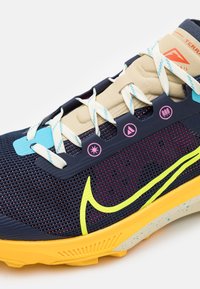 Marineblauwe en beige hardloopschoen met gele zool, neongele swoosh-logo, lichtblauwe accenten, witte veters met turquoise strepen en roze iconen op de zijkant.