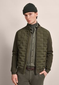 Olive green puffer jacket with a textured surface, paired with a matching turtleneck and button-up, complemented by brown belt and khaki pants.