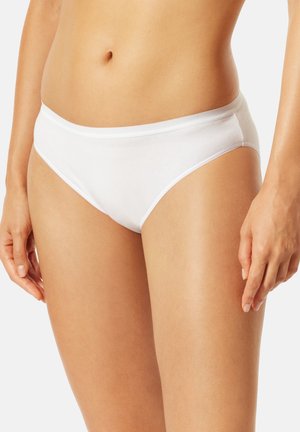 Frau trägt schlichte weiße Baumwoll-Unterwäsche im Bikini-Stil, die auf Höhe der Taille und bis zur Mitte des Oberschenkels beschnitten ist, steht vor einem schlichten hellen Hintergrund.