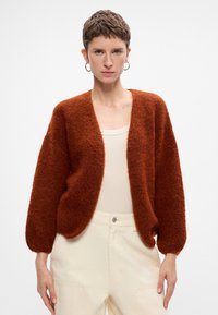 Brunrustfarvet fluffy cardigan med åben front, brede ærmer og blød tekstur, båret over en lys tanktop og cremefarvede bukser.