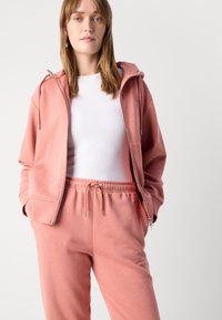Lichtroze hoodie en bijpassende joggingsbroek van zachte stof. De hoodie heeft een ritssluiting aan de voorkant, een trekkoord en zijzakken voor extra gemak.