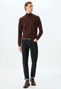 Brauner Rollkragenpullover, schmal geschnitten, kombiniert mit dunklen Jeans und braunen Stiefeletten. Die Jeans haben hochgekrempelte Säume und einen kontrastierenden Gürtel.