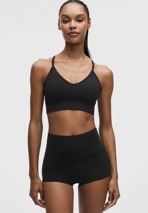 Kvinde stående iført en sort sports-bh med tynde stropper og matchende højtaljede atletikshorts mod en ensfarvet baggrund.