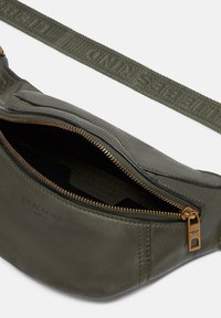 Olivgrüne Leder-Umhängetasche mit strukturierter Oberfläche, goldfarbenem Reißverschluss und einem gemusterten Stoffgurt, der den Markennamen zeigt.