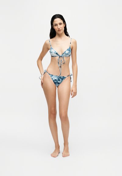 Femme aux cheveux foncés debout pieds nus portant un bikini tie-dye bleu et blanc avec fines bretelles et liens latéraux sur fond clair uni.
