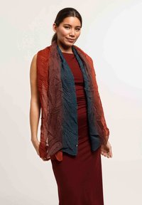 Écharpe légère arborant un motif de feuilles texturé, dégradé du rouge au bleu. Drapée sur une robe moulante bordeaux.