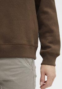 Brown sweatshirt with ribbed cuffs and hem, featuring a textured fabric. Paired with light gray pants, showing a hand resting at the side.