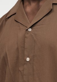 Chemise marron avec un col cranté et deux boutons blancs attachés, portée par une personne à la carnation medium-foncée.