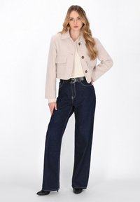 Kvinde med langt blond hår iført en beige cropped jakke, cremet sweater, brede mørkeblå jeans, sort bælte og sorte hælede støvler.