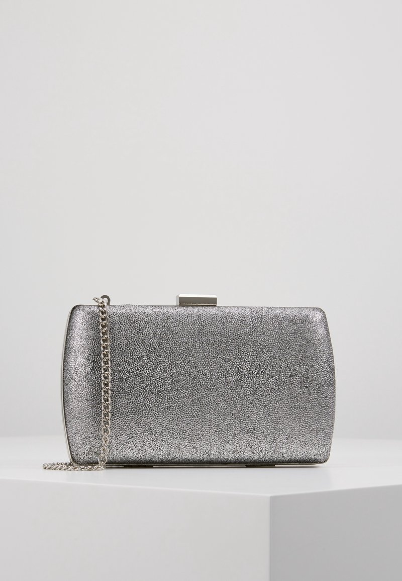 Pochette rectangulaire argentée avec surface texturée et bandoulière chaîne argentée sur un socle blanc sur fond gris uni.