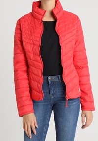 Rød quiltet jakke med høj krave, lynlås lukning og ribstrikket tekstur. Kombineret med en sort top og blå jeans.