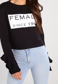 Svart kort sweatshirt med "FEMALE SINCE 198" tryckt i vitt. Har volangärmar och är kombinerad med ljusblå denimjeans.