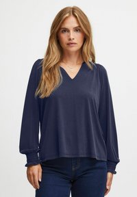Mörkblå långärmad topp med V-ringning, smockade ärmslut, avslappnad passform och mjukt material. Bärs tillsammans med mörka denimjeans.
