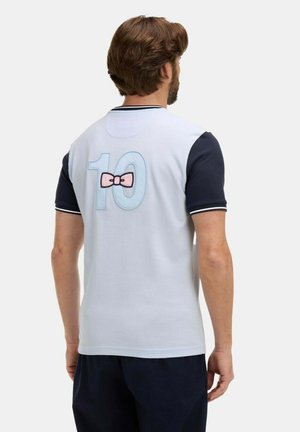 Homme aux cheveux bruns portant un t-shirt blanc et marine à manches courtes avec le numéro 10 et un motif de nœud rose dans le dos.