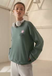Sudadera gris-verde con un gráfico de estrella rosa y el texto "DANCE PALACE". El modelo lleva auriculares blancos de diadema. Mangas largas, ajuste casual.
