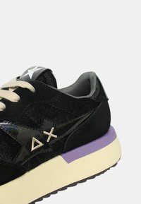 Baskets en suede noir avec des accents en vernis irisé, semelle crème, talon violet, doublure grise et lacets beiges. Logo présent sur la languette.
