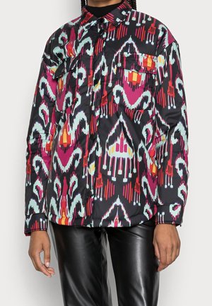 Osoba ubrana w zapinaną na guziki koszulę z długim rękawem w kolorowy, abstrakcyjny wzór geometryczny oraz czarne spodnie z ekoskóry.