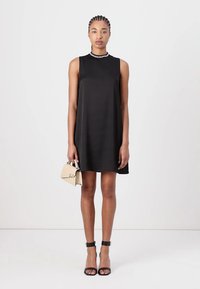 Black sleeveless dress with a fitted bodice, flared skirt, and smooth texture. Paired with black ankle strap heels and a small beige handbag.