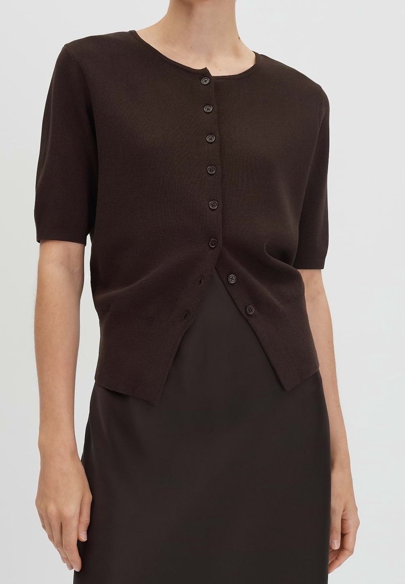 Femme portant un cardigan boutonné à manches courtes brun foncé sur une robe brun foncé devant un fond clair uni.