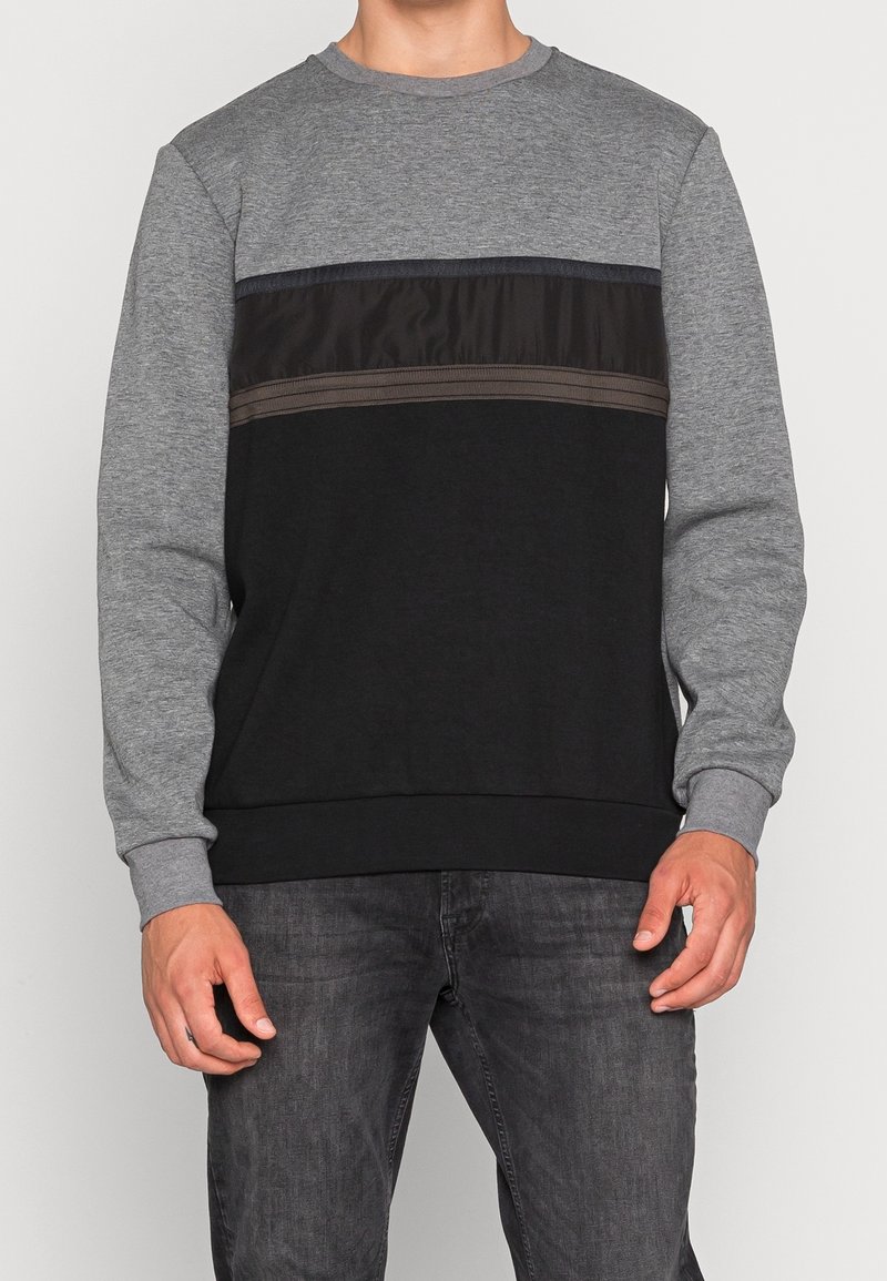 Homme portant un sweat-shirt à manches longues avec un col rond, des manches grises et un corps noir avec des rayures horizontales marron foncé et noires.