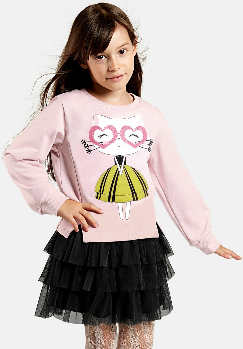 Svetlo roza pulover s grafiko mačke, ki nosi očala v obliki srca, v kombinaciji z črno slojevito tila krilom in belimi pajkicami s vzorcem.