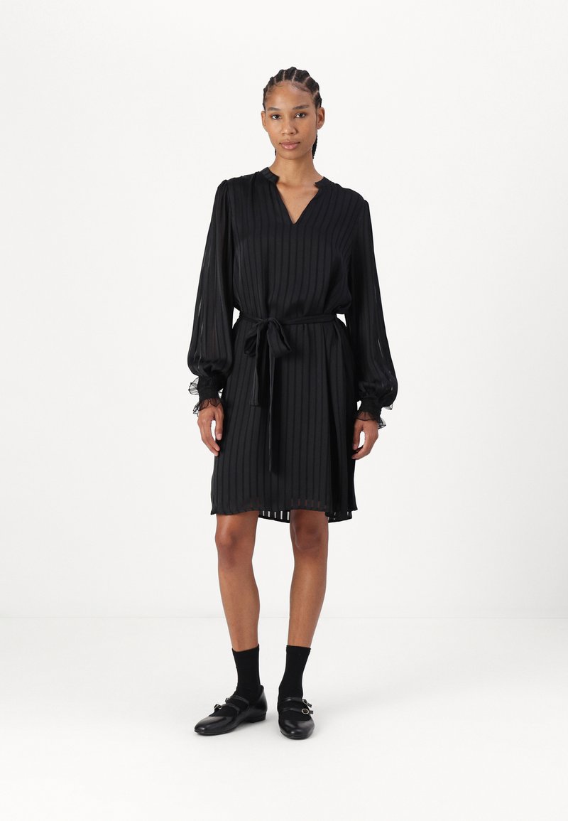 Črna nabrana obleka z V-izrezom, dolgimi rokavi in pasom na zavezovanje. Ima nabrane manšete in zračno silhueto. Nosijo se z črnimi nogavicami in čevlji.