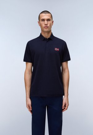 Man met kort haar die een marineblauw poloshirt draagt met een klein Noors vlagembleem op de borst, staand tegen een effen achtergrond.