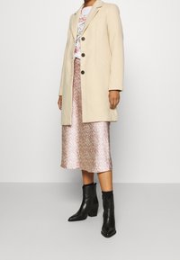 Cappotto in misto lana beige con colletto a rever, tre bottoni, indossato sopra una gonna midi di satin floreale rosa e stivaletti neri con tacco spesso.