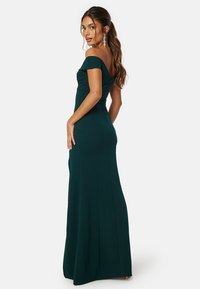 Abito verde smeraldo off-shoulder con corpetto aderente, orlo svasato e dettagli di cucitura in vista. Tessuto lungo e liscio con silhouette elegante.