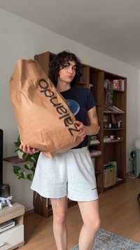 Eine Person mit lockigem Haar, die ein dunkelblaues Shirt und graue Shorts trägt, hält eine große braune Zalando-Einkauftasche in einem Wohnzimmer mit Regalen aus Holz.