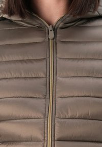 Close-up van een bruine gewatteerde jas met een gouden rits, gedragen door een persoon met kort bruin haar.