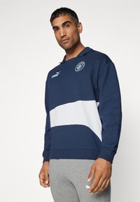 Sweatshirt bleu marin cu o bandă orizontală albă, guler polo și logo Puma. Manșete cu lână pe mâneci lungi și un emblema rotundă pe piept.