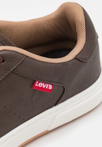 Brune lærersneaker med en tan mesh krage, hvit gummisåle og liten rød Levi's-logo på siden. Teksturert finish detaljer til stede.