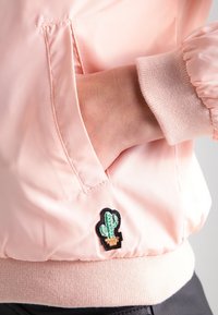 Różowa kurtka bomber z błyszczącym wykończeniem, ściągaczami na mankietach oraz małą czarno-zielono-beżową naszywką kaktusa na przedniej kieszeni.