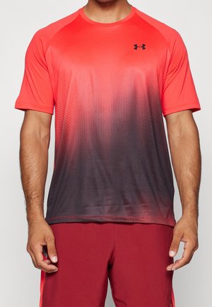 Mężczyzna w czerwono-czarnym koszulce sportowej Under Armour z krótkim rękawem o gradientowym wzorze oraz czerwonych szortach, stojący z rozluźnionymi ramionami.