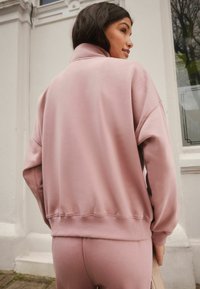 Felpa oversize rosa realizzata in tessuto morbido; presenta un colletto alto e orlo a coste. Il modello indossa leggings rosa abbinati.