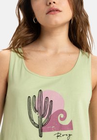 Ljusgrön ärmlös tanktop med en kaktus- och vågdesign i svart och rosa. Mjuk tyger med rund halsringning och avslappnad passform.